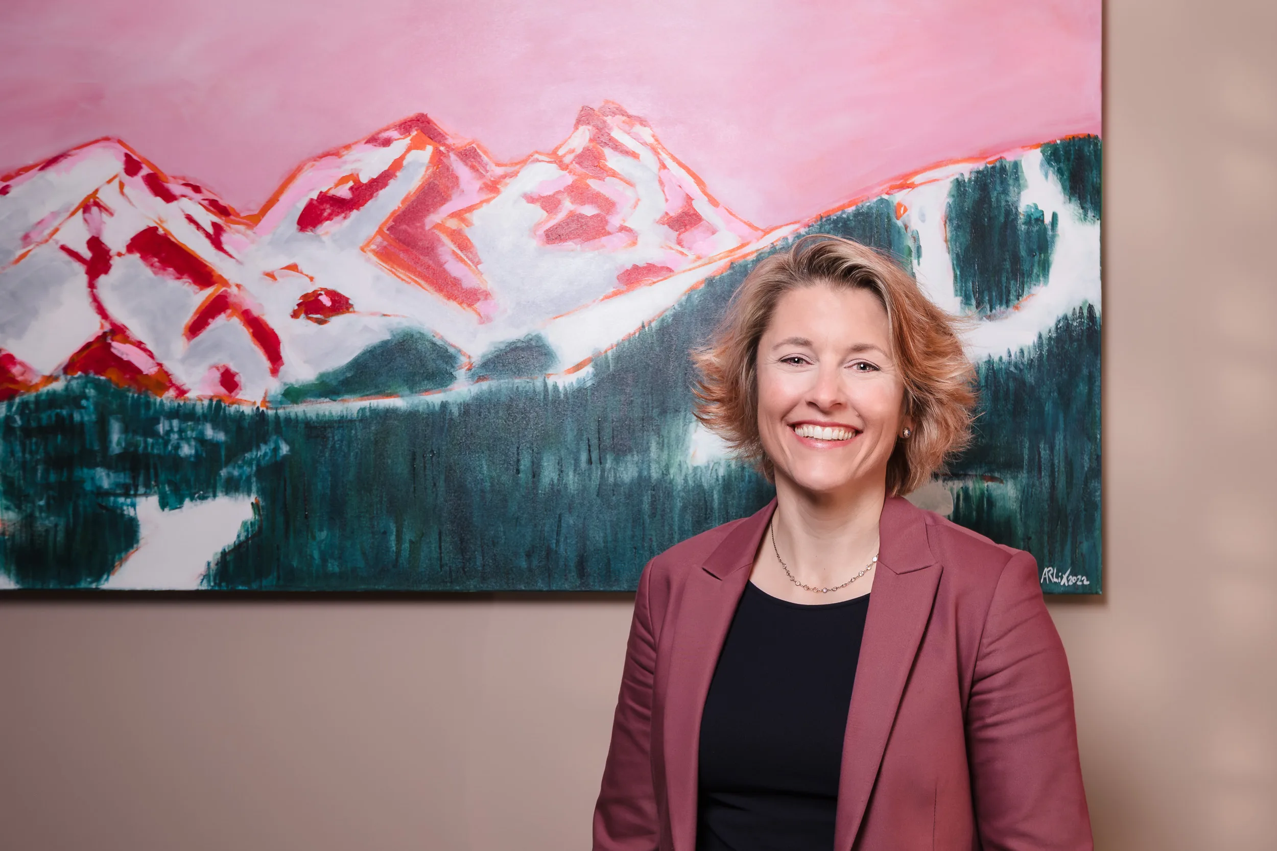 Frau mit passendem roten Blazer lacht vor einem Berg-Gemälde.